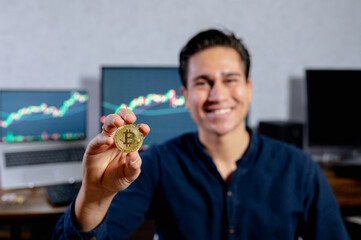 Young smiling hispanic man showing a bitcoin cash coin. computer with. Computers showing growth charts of value currency on background.