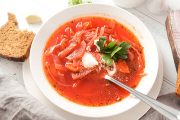 borscht with sour cream and herbs in a white plate,  bread, spoon on a  wooden table top view