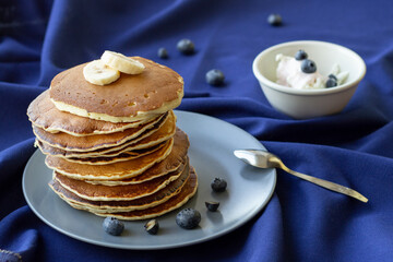 Stack of pancakes on a plate with bananas and blueberries on blue tablecloth