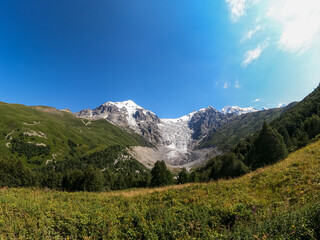 Fototapeta premium A panoramic view on the snow-capped peaks of Tetnuldi, Gistola, Lakutsia and the Adishi Glacier in the Greater Caucasus Mountain Range in Georgia, Svaneti Region. Sharp peaks, wanderlust, solitude.