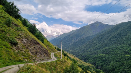 Fototapeta premium A mountain road in the Greater Caucasus Mountain Range in Georgia, Svaneti Region. Electricity lines are built up along the valley. In the background are sharp snow-capped mountains. Adventure,Freedom
