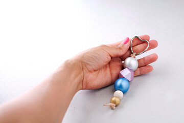 Female hand holding keychain of painted wooden beads with heart-shaped ring on a light background. Wooden beads and hexagon in pink, green, blue and metallic gold colors. Handmade and crafts concept.