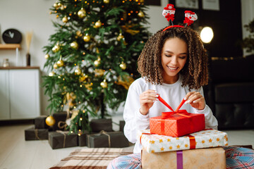 Fototapeta na wymiar Afro American woman in pajamas and in santa claus hat with large gift box posing near Christmas tree. Winter holiday, vacation, relax and lifestyle concept.
