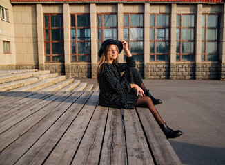 A young beautiful girl in a dress and hat is sitting on a wooden podium.There are beautiful vintage windows.