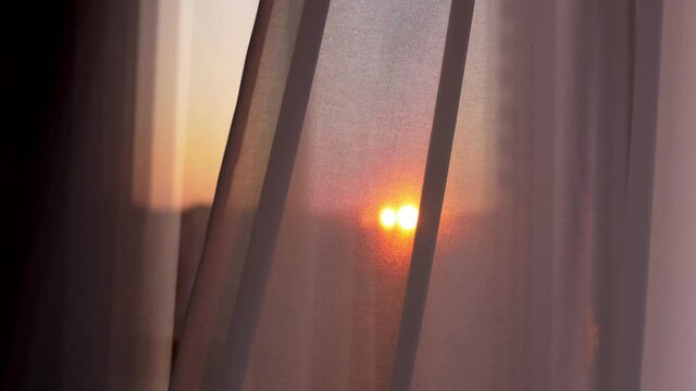 The window is covered with a white tulle curtain. Tulle curtains add coziness and comfort to the home. The kind sunlight of the setting sun seeps through the tulle curtain. The tulle sways gently.
