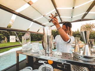 cheerful bartender woman mixing drinks and cocktails outdoors near a pool on a park of a venue...