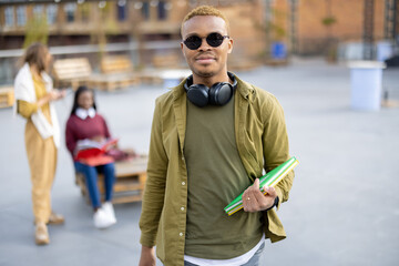 Smiling black student guy on background of her female friends outdoors. Concept of education and learning. Idea of student lifestyle. Young man holding books and looking at camera. University campus