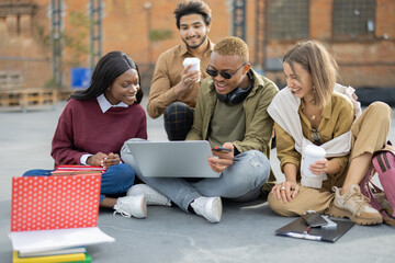 Multiracial students sitting and watching something on laptop computer on asphalt at university campus. Concept of education. Remote and e-learning. Idea of student lifestyle. Young smiling friends