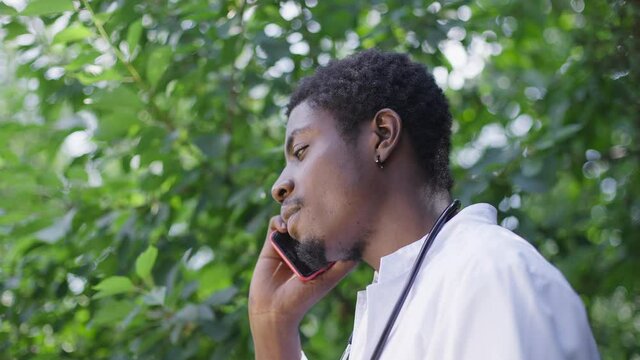 Young doctor standing outdoors on sunny hospital backyard talking on phone in slow motion. African American man on break outdoors with smartphone. Lifestyle and medicine concept