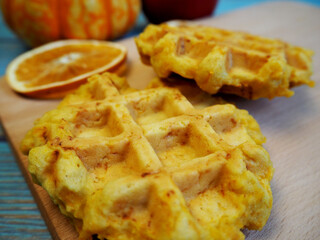 close up, gluten-free and lactose-free Belgian pumpkin waffles lie on a wooden board with a dry slice of lemon next to a small pumpkin and an apple on a blue wooden table background. side view