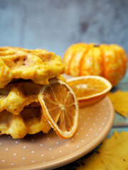 pumpkin Belgian waffles lie on a brown plate with two dry slices of lemon on a background of yellow maple leaves on a blue table and a small pumpkin at the back. top view. gluten-free and lactose-free