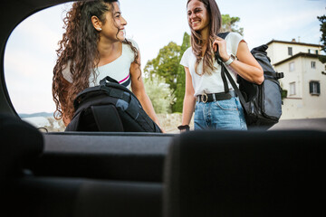 Portrait of two young female friends from inside car as take backpacks for the hike during vacation...