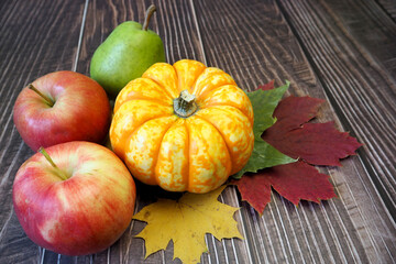 a small yellow pumpkin, two red apples and a green pear lie on yellow and red maple leaves on a wooden background. side view