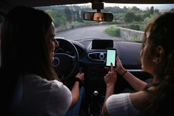 Two young women in a car on a road trip in the evening at sunset check the map on smartphone to geo...