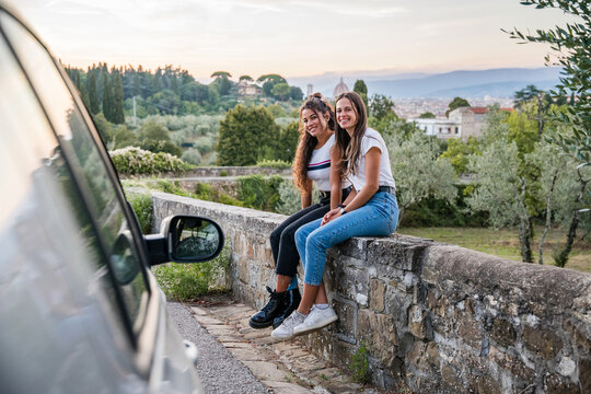 Portrait of two young women sitting on the low wall at sunset in a car trip among nature, behind them the city and vegetation - Millennials having fun together during an unforgettable vacation