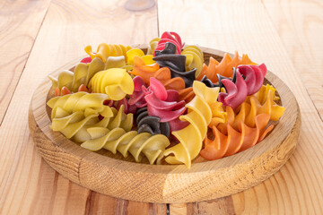 Uncooked colored fusilli pasta in a wooden dish on a wooden table, close-up.
