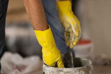 Hold a spatula in your hands and dilute the plaster in a bucket. The hands are wearing yellow gloves. The builder makes repairs in the apartment.