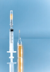 A syringe with a burning needle and divisions next to a glass ampoule with a gold glowing filling, vertically on a blue background.