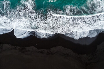 Overhead view of a beach with black sand and the blue water of the sea.