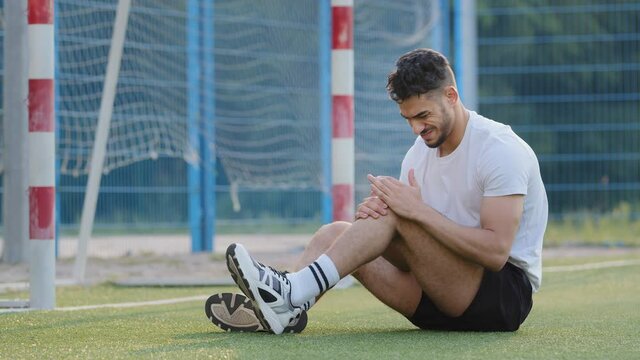 Unhappy injured Middle Eastern footballer sitting on grass of soccer field against goal, holding knee. Millennial athlete in sportswear suffers from leg pain experiencing about injury, cannot continue
