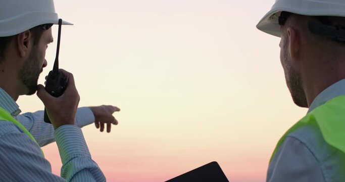 Employees in protective helmets and reflective vests regulate ships loading by dockside cranes via tablet and radio set at sunset closeup