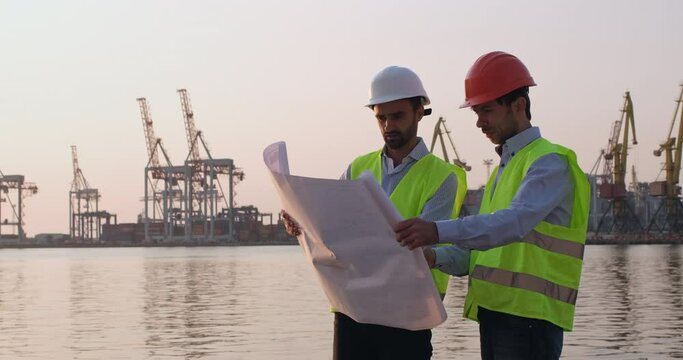 Engineers wearing helmets and reflective vests regulate building of cargo cranes using delineation standing on beach against port