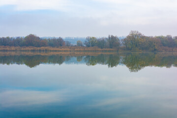 Cold foggy river in the morning with overgrown banks and displaying clouds in the water.