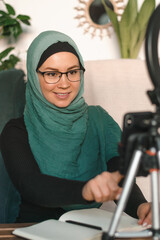 A young Muslim woman in a hijab leads her video blog, live broadcast.Cozy home interior with indoor plants.Social media and technology concept,diverse people.Selective focus.