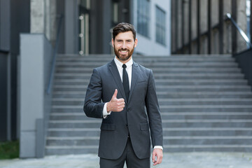 Portrait of young successful businessman looking at camera and smiling outdoors holding thumbs up...