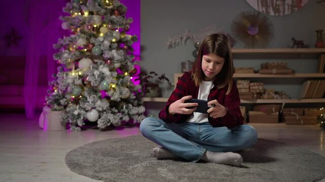 Cute long-haired teenage guy playing in a smartphone on New Year's Eve, sitting on a carpet waiting for the celebration in a festive interior. Living room with decorated Christmas tree with gift boxes