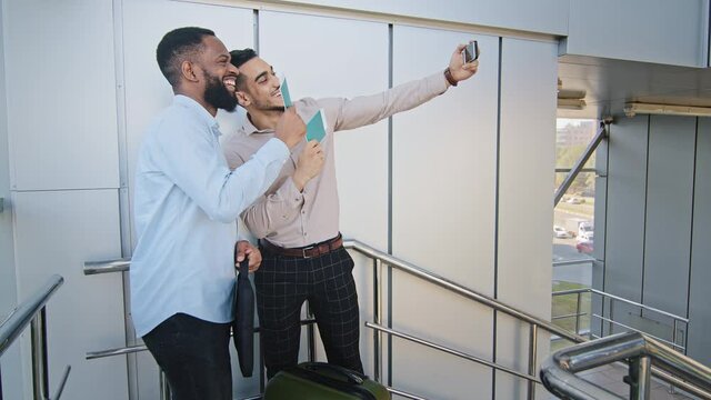 Two multiracial friends business partners happy hispanic man and smiling african guy with suitcase standing in airport taking selfie photo using mobile phone showing passports plane tickets video call