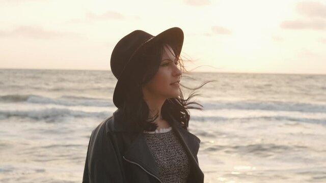 Portrait of romantic brunette woman in hat and coat walking on the beach in autumn at sunset or sunrise 