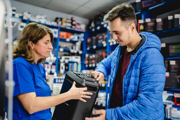 Front view portrait of young adult caucasian man standing by female seller in the electronics store looking and peaking products checking wireless speaker wearing blue jacket real people copy space