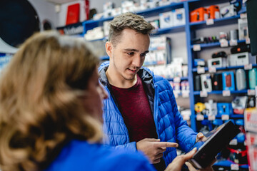 Front view portrait of young adult caucasian man standing by female seller in the electronics store looking and peaking products checking wireless keyboard wearing blue jacket real people copy space - Powered by Adobe
