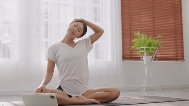 In the fitness studio, an Asian woman is studying online fitness training. Improve your health by extending your neck and meditating.