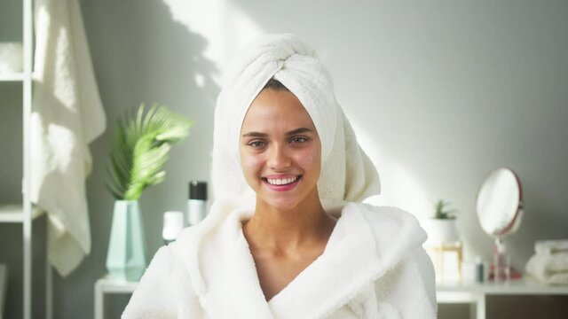Happy woman smiling in bathroom or spa salon. Portrait of young female student wearing white bathrobe and towel on head after shower. Natural day make up, facial skin care concept.