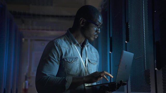 Engineer serving of contemporary data center. African-American person with glasses spbas illuminated by laptop screen sets up powerful servers at racks closeup