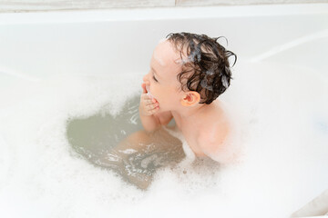 A small child takes a bubble bath in a beautiful bathroom. Shampoo, hair care product and soap for children. The child is bathing in a large tub. A little boy with foam in his hair.