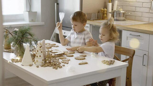 Boy and girl decorate and eating Christmas gingerbread at home. A boy and a girl paint with cornets with sugar icing on cookies. New Year's decor, branches of a Christmas tree.