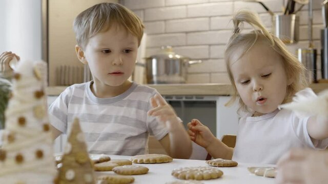Mom and children decorate Christmas gingerbread at home. A boy and a girl paint with cornets with sugar icing on cookies. New Year's kitchen decorations, branches of a Christmas tree.