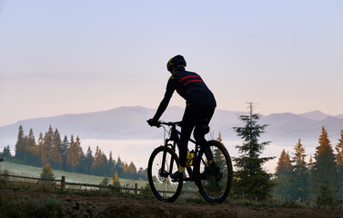 Fototapeta premium Silhouette of cyclist riding bike with coniferous trees and hills on background. Back view of man bicyclist enjoying bicycle ride in mountains in the morning. Concept of sport and active leisure.