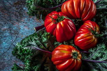 Flat lay of big fresh ripe tomatoes with kale leaves and wooden cutting board on the rustic dark blue background