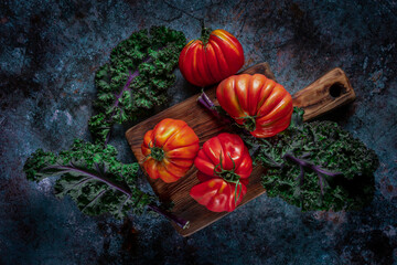 Flat lay of big fresh ripe tomatoes with kale leaves and wooden cutting board on the rustic dark blue background