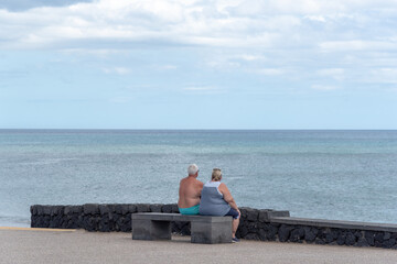 un pareja de personas mayores de espaldas sentado en un banco mirando el mar cerca de la playa. 