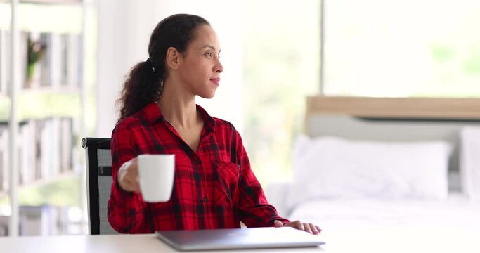 Cheerful ethnic woman in casual clothes closing laptop and taking mug of hot beverage while sitting at desk and working on remote project at home