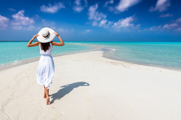 Fototapeta na wymiar A woman in a white summer dress walks on a tropical paradise beach with turquoise sea and sunshine