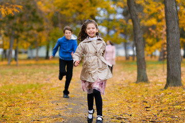 Fototapeta premium Children run along path in autumn park. They play and laugh, they have a lot of fun. Beautiful nature and trees with yellow leaves.
