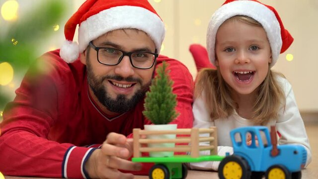 Toy tractor delivers Christmas tree in cart to happy family in red santa hats. Father gives little daughter new year gift.