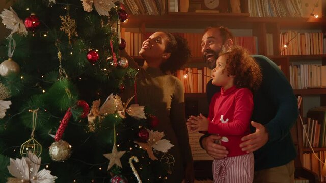 Preschool girl and her loving parents decorating Christmas tree