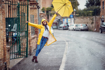 A young girl with yellow raincoat and umbrella is having a good time while walking the city on the rain. Walk, rain, city - obrazy, fototapety, plakaty